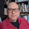 Headshot of Richard Hill FAHACS MA student sat in front of a bookshelf. Richard wears a black t shirt and red fleece.